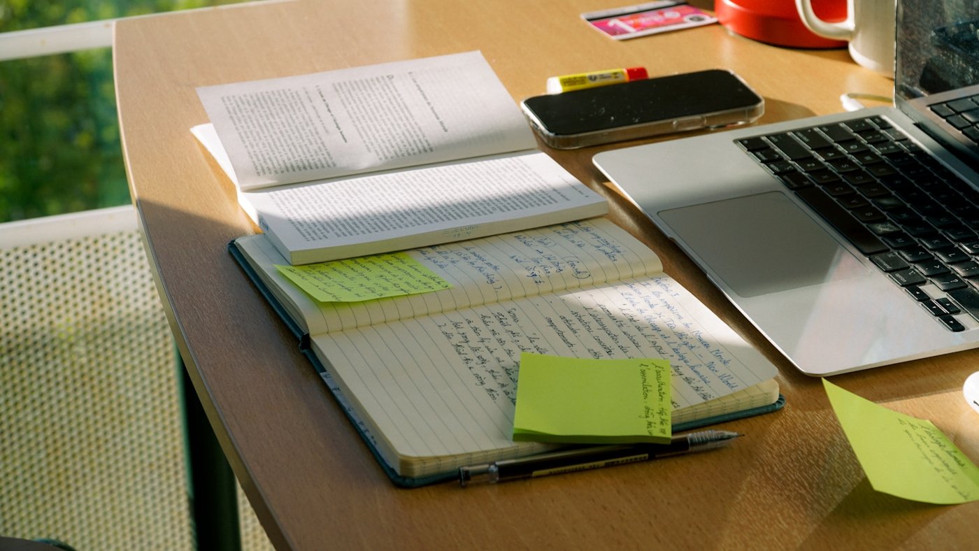 A cluttered desk with a laptop, notebook, and scattered notes